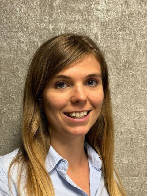 Person with long, light brown hair wearing a light blue button‑down shirt, standing against a textured gray wall.
